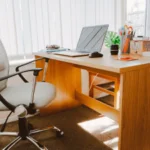 Alt tag: A sunlit home office featuring a wooden desk with an open laptop, a black ergonomic chair, stationery holders, and a small potted plant, with vertical blinds partially closed in the background.