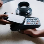 Person making a contactless payment with a smartphone over a card reader with a cup of coffee in the background on a table.