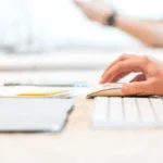 Person working on a computer with documents and a tablet on the desk.