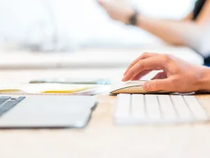 Person working on a computer with documents and a tablet on the desk.