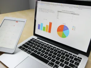 A workspace featuring an open laptop displaying colorful bar graphs and pie chart on the screen, alongside a tablet showing a monthly calendar.