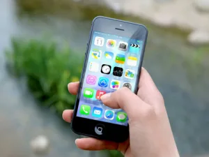 A hand holding a smartphone with colorful app icons on the screen, blurred greenery in the background.