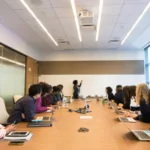 Alt text: A modern conference room setting with a diverse group of professionals seated at a long table, attentively facing a presenter at the whiteboard, with laptops and notebooks on the table indicating an active meeting or training session in progress.