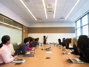 Alt text: A modern conference room setting with a diverse group of professionals seated at a long table, attentively facing a presenter at the whiteboard, with laptops and notebooks on the table indicating an active meeting or training session in progress.