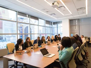 A diverse group of professionals engaged in a meeting around a conference table with laptops in a modern office setting.