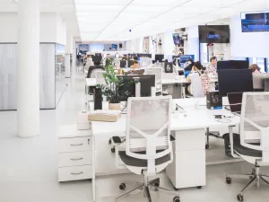 Alt text: Modern office space featuring rows of white desks with computers, ergonomic chairs, and individuals working, accented with green plants and natural light.