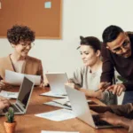 A diverse group of professionals collaboratively working around a table with laptops and documents in a brightly lit office setting.