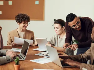 A diverse group of professionals collaboratively working around a table with laptops and documents in a brightly lit office setting.