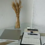 A modern home office setup with a closed laptop, a clear clipboard with papers, glasses, smartphone, pen, and a vase of dried wheat on a glass table against a white brick wall.