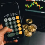 Hand holding a smartphone with a calculator app displayed, showing a large number, with a blurry background featuring a laptop screen with stock market graphs and physical coins.