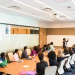 Alt text: A diverse group of professionals attending a presentation by a speaker in a well-lit conference room with large windows.