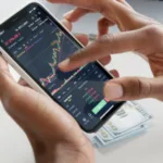 A person's hands holding a smartphone displaying stock market charts, with US dollar bills lying underneath on a table.