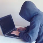 Person in a gray hoodie typing on a laptop with code on the screen, suggesting a programmer or hacker at work, in a room with a white backdrop.