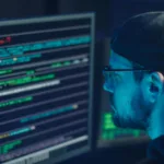 Man wearing glasses and cap focused on code on computer monitors in dimly lit room, reflecting cyber work environment.