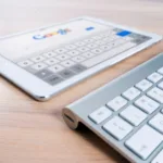 A tablet with a Google search page displayed lies on a wooden desk next to a white Bluetooth keyboard, emphasizing modern wireless technology and internet access.