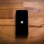 Alt text: A black iPhone with a lit Apple logo centered on a wooden parquet surface.