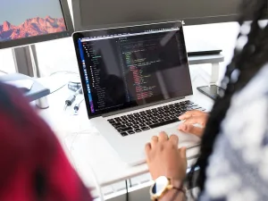 Person working on a laptop with code on the screen, sitting at a desk with a mountain view outside the window.