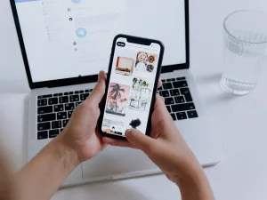 Person holding a smartphone with Pinterest on the screen in front of an open laptop on a white desk.