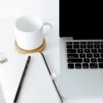 A clean and simple workspace featuring an open notebook with a pen on top, a white mug on a coaster, and a laptop with black keys on a white desk.