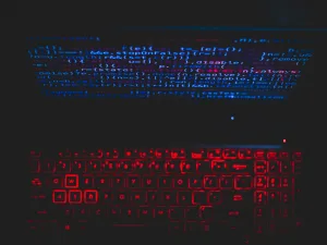 Alt text: Backlit keyboard with red illumination against a dark background, with the glow of coding text on a computer screen above.