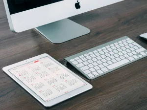 An Apple iMac with keyboard on a wooden desk, alongside an iPad displaying a calendar.