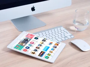 An Apple iMac on a desk with a keyboard, a mouse, a glass of water, and an iPad displaying colorful app icons.