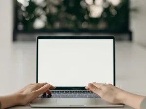 Person typing on a laptop with a blank white screen, set on a table with a blurred background of indoor plants.