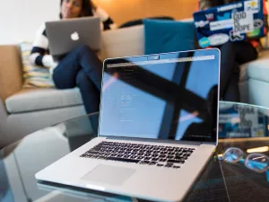 An open laptop with code on the screen in focus on a glass table with a blurred person working in the background.