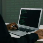 Alt text: A person coding on a laptop with a screen displaying lines of code, set against a dark backdrop with out-of-focus digital data.