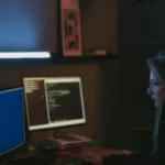 A person working intently on a computer with multiple screens in a dark room, illuminated by the soft glow of the monitors.