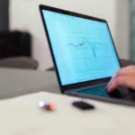 Close-up of a person's hand using a laptop displaying graphs, with a smartphone and wireless earbuds on the table.