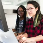 Two women collaboratively working at a computer in an office environment.