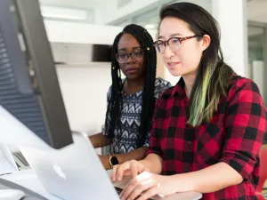 Two women collaboratively working at a computer in an office environment.