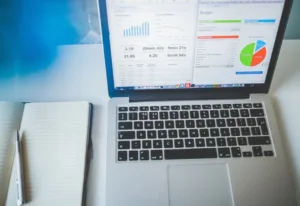 Workstation with an open laptop displaying graphs and charts on the screen, alongside a notebook with a pen on a white desk.