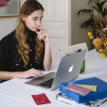 Woman in black shirt working on a MacBook at a white desk with a stack of books and a vase of flowers.