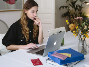 Woman in black shirt working on a MacBook at a white desk with a stack of books and a vase of flowers.